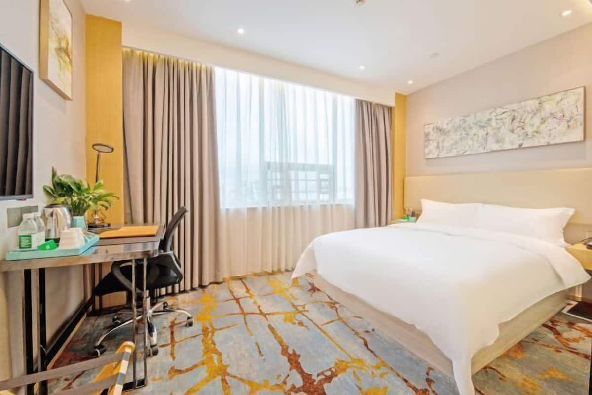 A hotel room with a large bed, a desk with a chair, a plant, and a window with curtains.