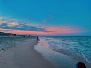 A beach with a calm sea, a clear sky, and a few people walking along the shore.