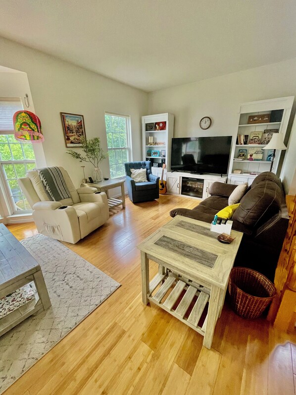 A living room with a sofa, armchair, coffee table, TV, bookshelf, and a window with a view of greenery.