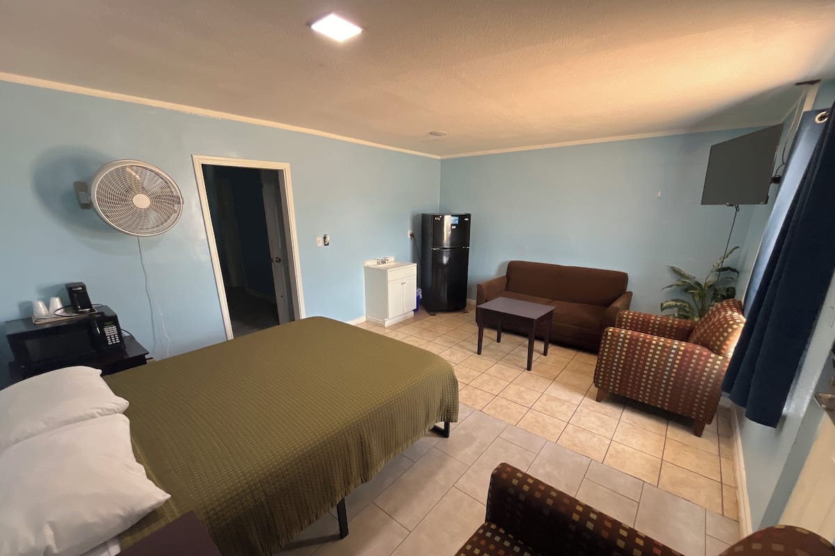 A hotel room with a bed, a sofa, a small table, a fan, a refrigerator, and a TV.