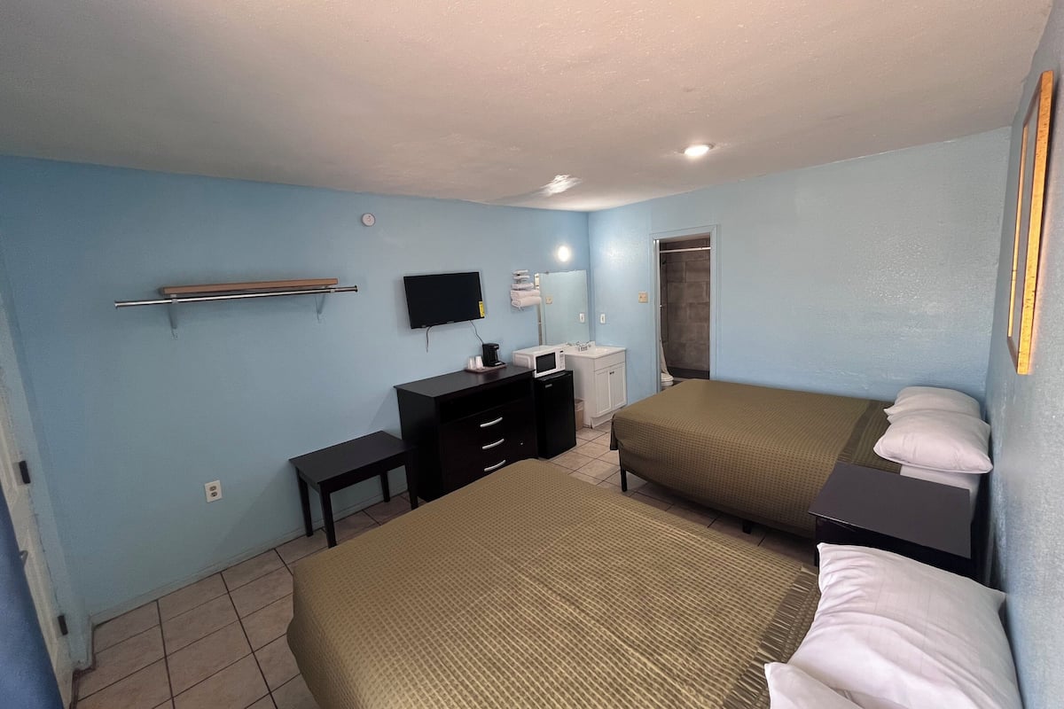 A hotel room with two beds, a desk, a TV, a small kitchenette, and a shower area.