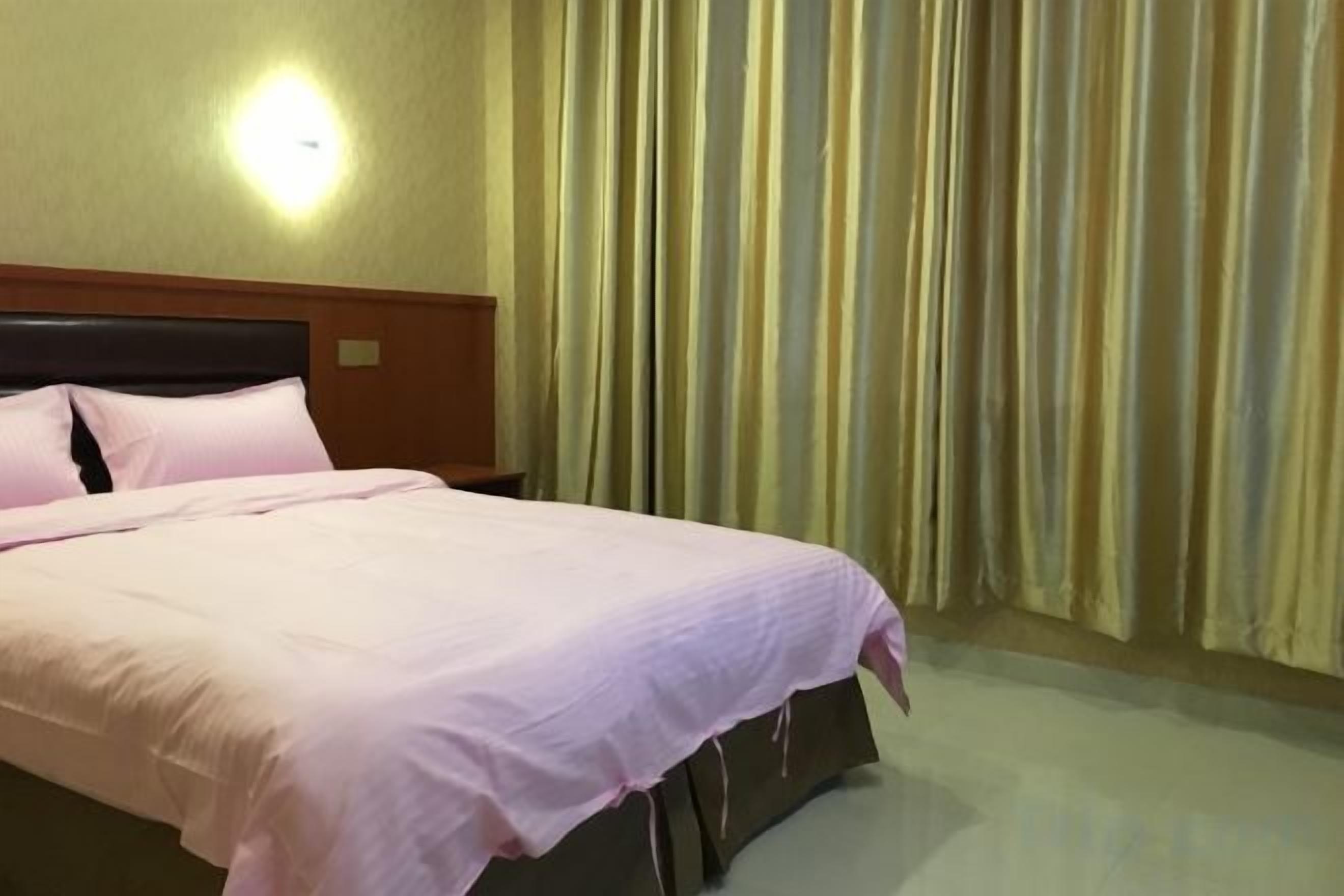 A hotel room with a bed, a headboard, a wall-mounted light, and a curtain.