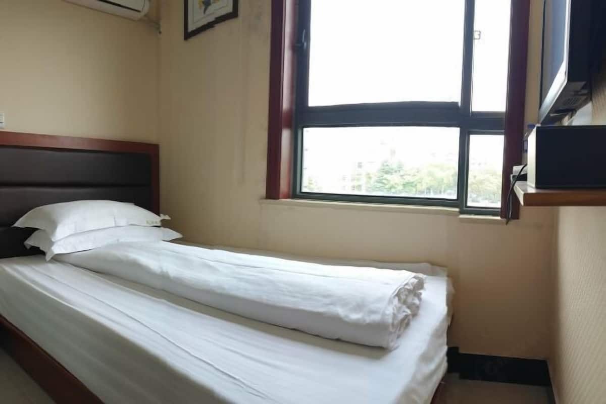 A single bed with white linens, a window with a view of buildings, and a wall-mounted air conditioning unit.