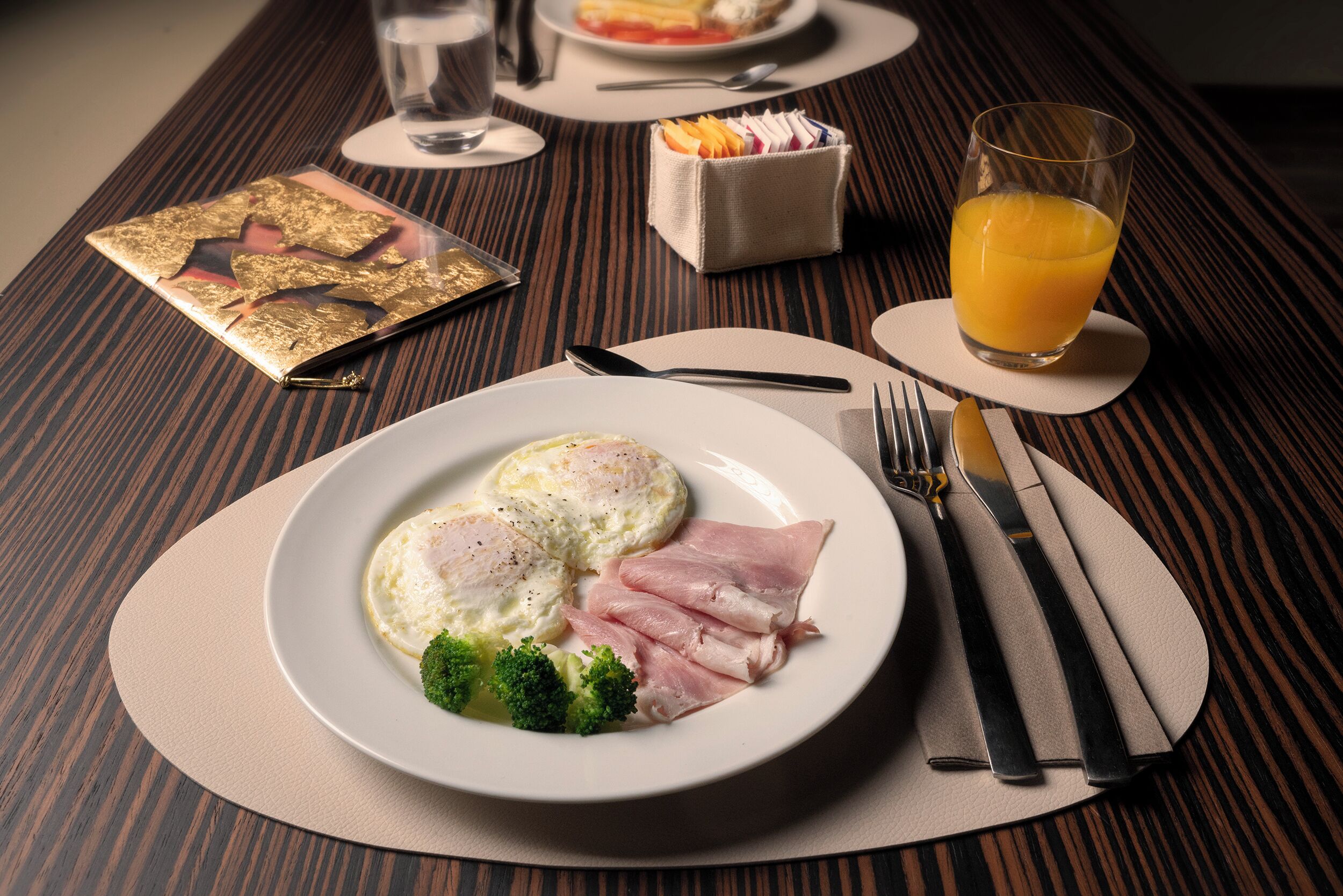 A breakfast setting with a plate of eggs, ham, and broccoli, a glass of orange juice, and a small container of condiments on a wooden table.