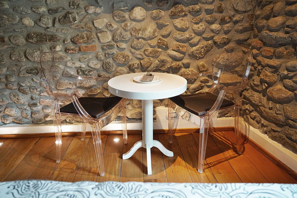 Una mesa redonda blanca con un plato y un tenedor, flanqueada por dos sillas transparentes con cojines negros, frente a una pared de piedra.