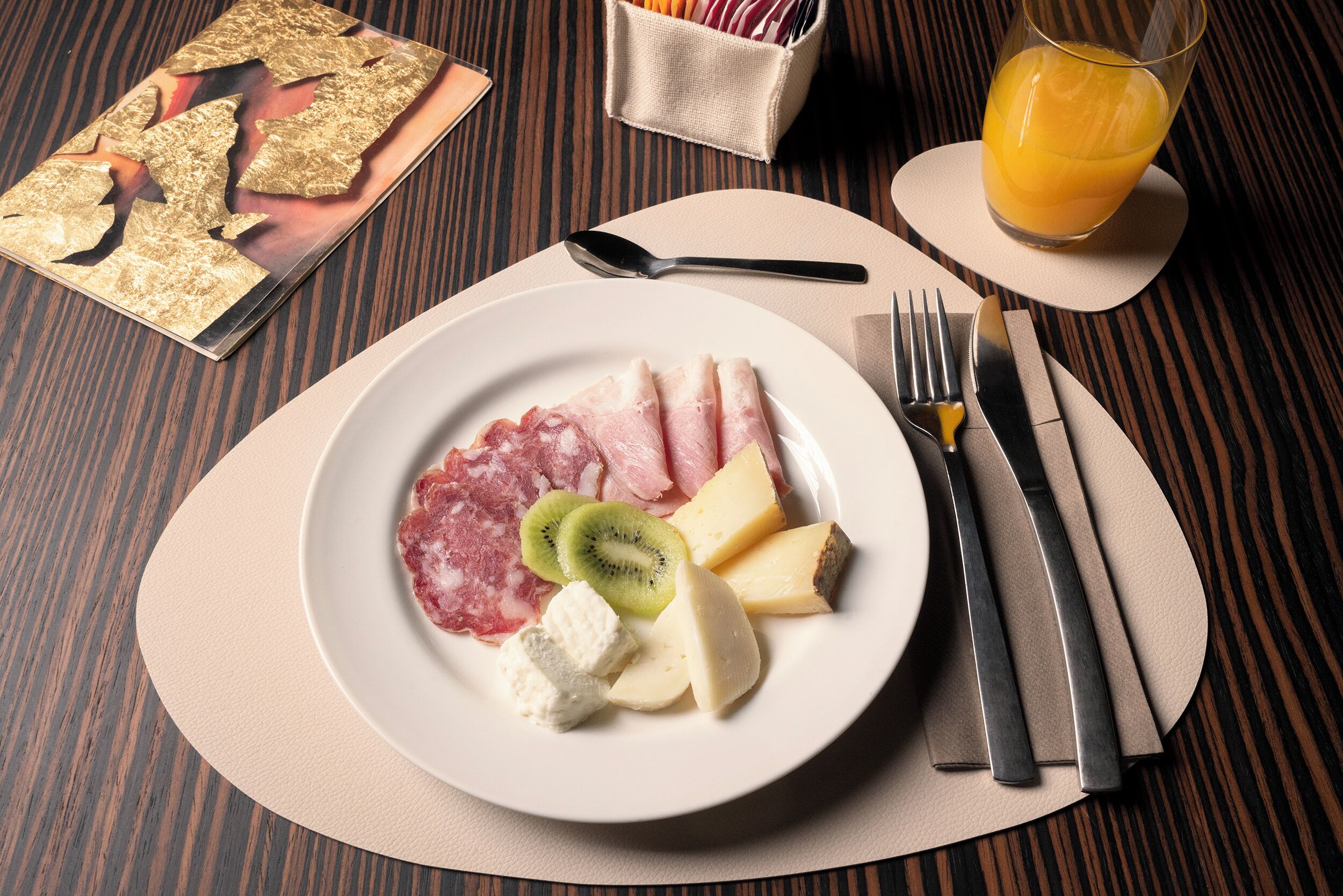A plate of food with ham, cheese, kiwi, and other items on a table with a spoon, fork, and knife.