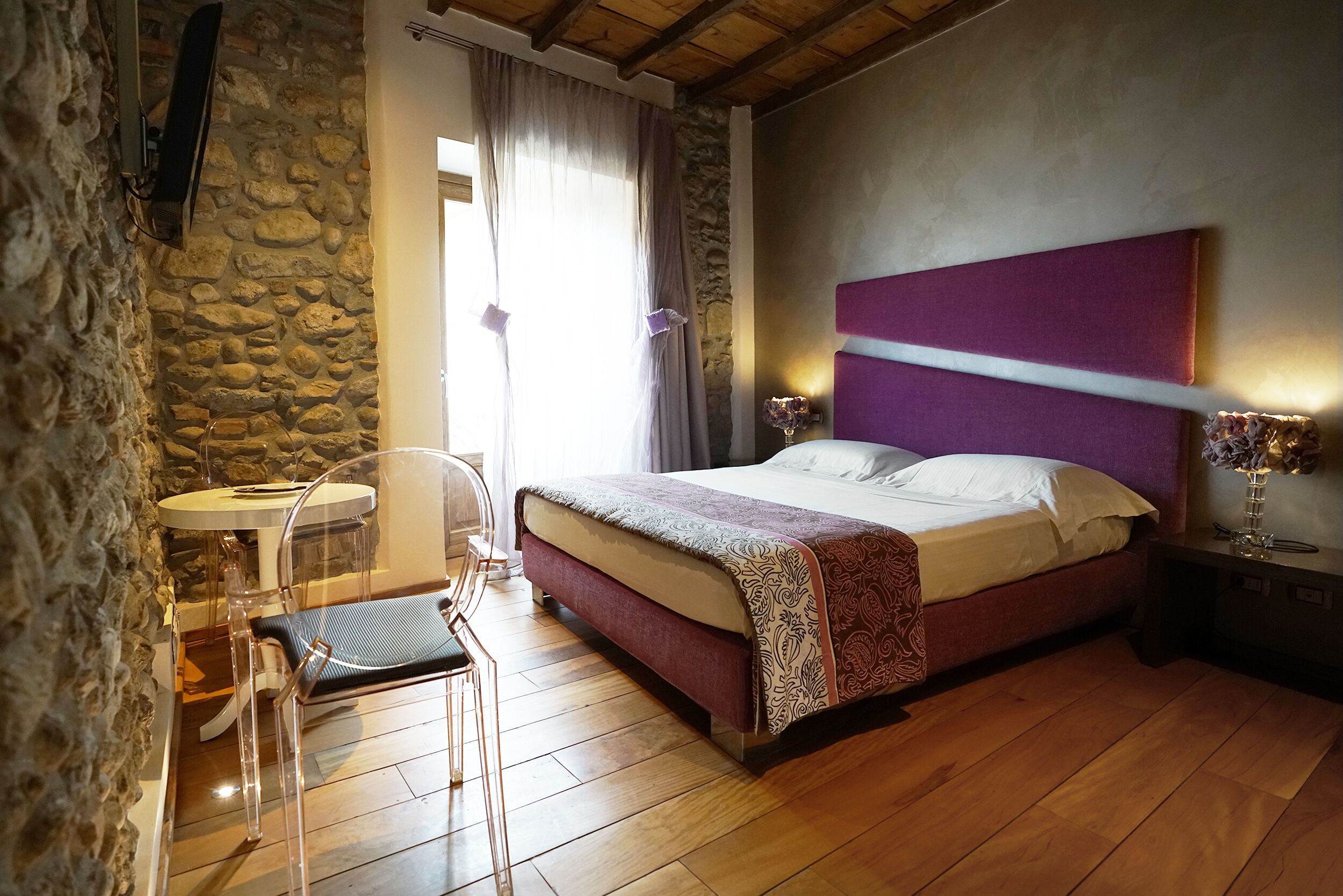 Ein Schlafzimmer mit Steinwand, einem Bett mit violettem Kopfteil, einem Nachttisch mit Lampe und einem kleinen runden Tisch mit Stuhl.