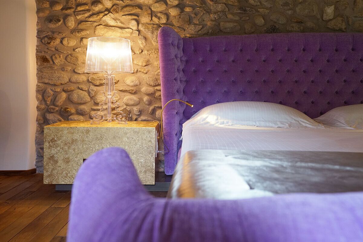 A bedroom with a stone wall, a purple upholstered headboard, a bedside table with a lamp, and a bed with white linens.
