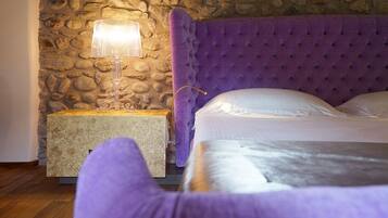 A bedroom with a stone wall, a purple upholstered headboard, a bedside table with a lamp, and a bed with white linens.