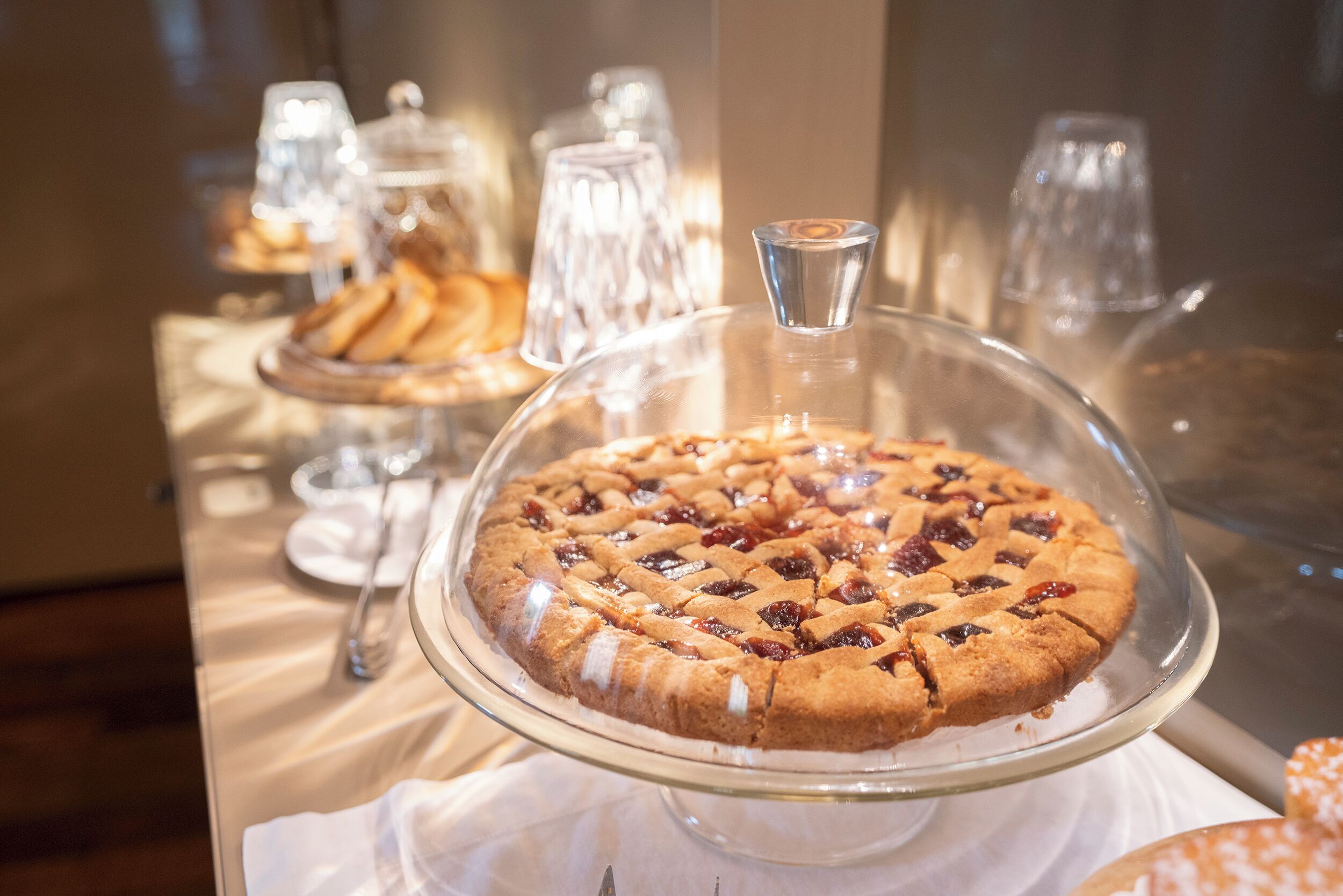 A dessert display with a covered pie on a glass stand, accompanied by a glass and a plate.