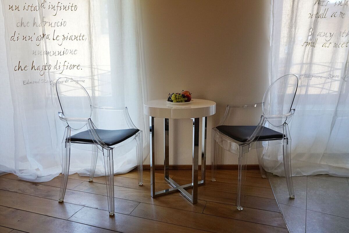 A room with two transparent chairs, a small white table with a fruit bowl, and a quote on the wall.