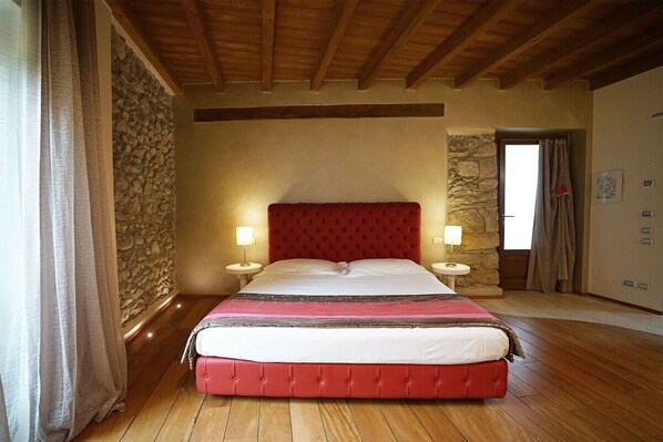 A bedroom with a red upholstered bed, stone wall, wooden floor, and a stone fireplace.