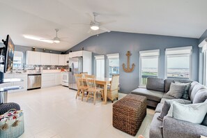 A spacious living area with a grey sofa and ottoman, a dining table with chairs, and a kitchen with white cabinets and a ceiling fan. - Propeller Clubhouse - Stunning Beach Views, Walk Steps to Beachfront (Galveston)