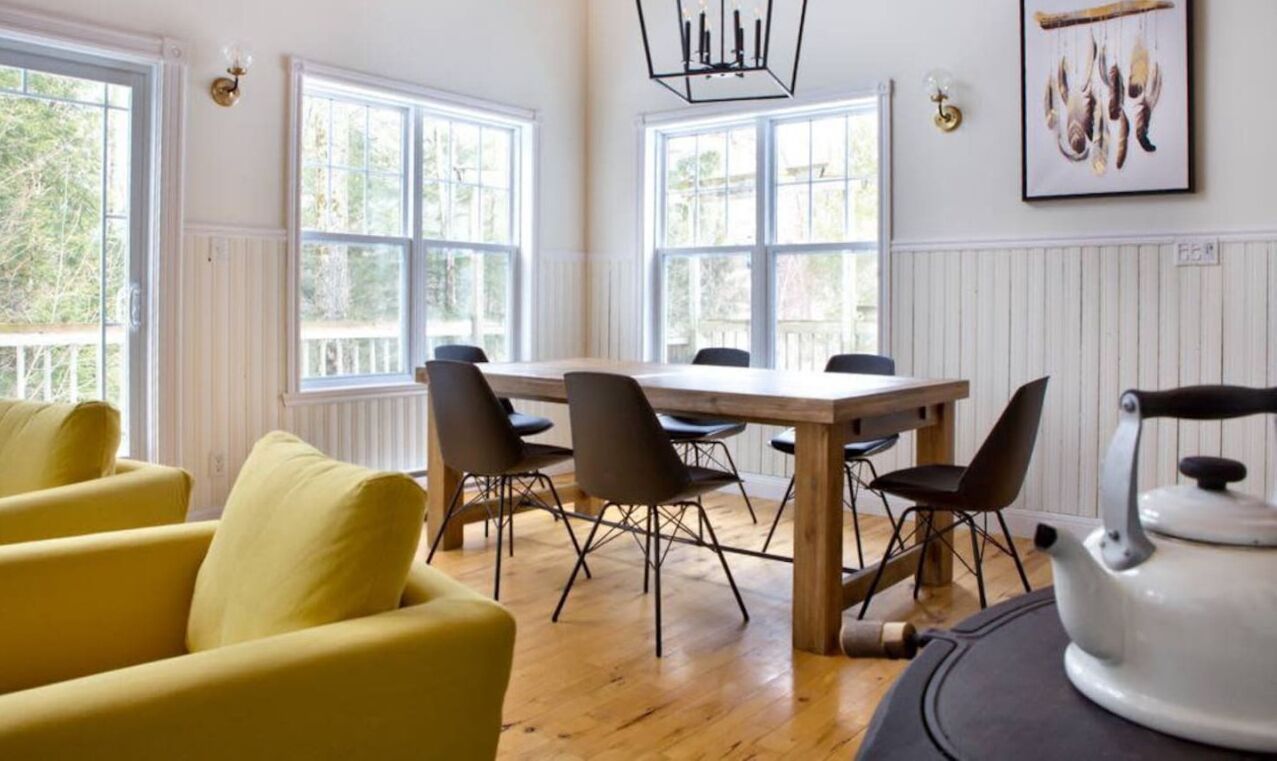 A dining area with a wooden table and chairs, large windows, and a yellow sofa.