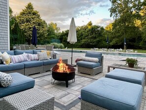 An outdoor patio with a fire pit, blue sofas, and a pool in the background.