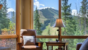 Ein gemütliches Zimmer mit Ledersessel, Holztisch und Lampe. Ein großes Fenster bietet Blick auf einen bewaldeten Berg.