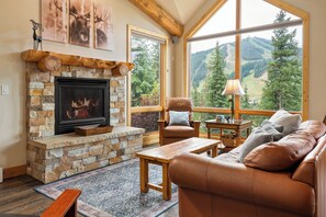 A cozy living room with a stone fireplace, leather sofa, and a large window offering a mountain view. - Alpine townhouse near Winter Park w/ hot tub (Winter Park)
