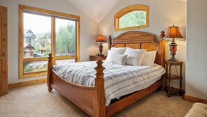 Ein Schlafzimmer mit großem Fenster, einem hölzernen Bett, zwei Nachttischlampen und Blick auf ein Haus und Bäume.