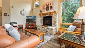 Ein gemütliches Wohnzimmer mit Kamin aus Stein, Ledermöbeln und einem großen Fenster mit Blick auf den Wald.
