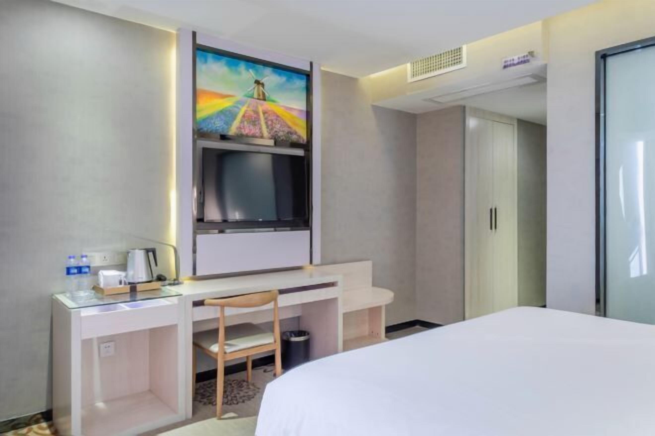 A hotel room with a bed, a desk with a chair, a television mounted on the wall, and a wardrobe.