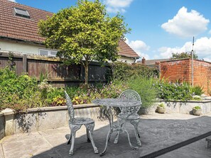 Eine Terrasse mit zwei verzierten Metallstühlen und einem Tisch, umgeben von einem Garten mit verschiedenen Pflanzen und Blumen.