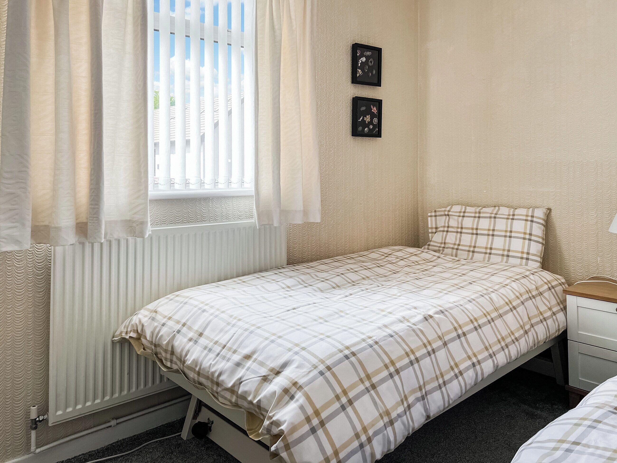 Ein Einzelbett mit karierter Bettdecke, ein Nachttisch mit Lampe und ein Fenster mit weißen Vorhängen.