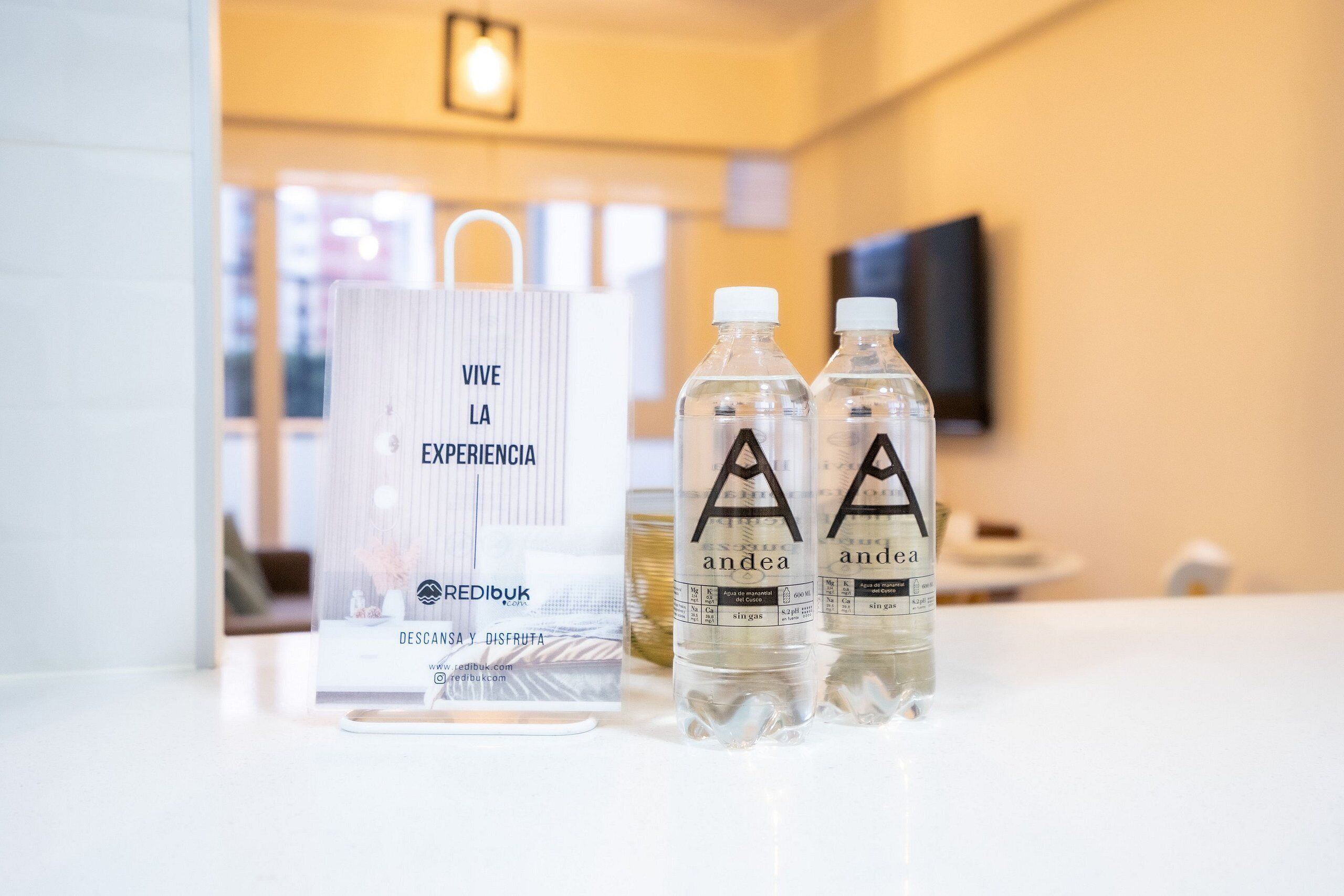 Zona de recepción de un hotel con un expositor de vidrio transparente, dos botellas de agua con la etiqueta 'andea' y un cartel blanco con el texto 'VIVE LA EXPERIENCIA'.