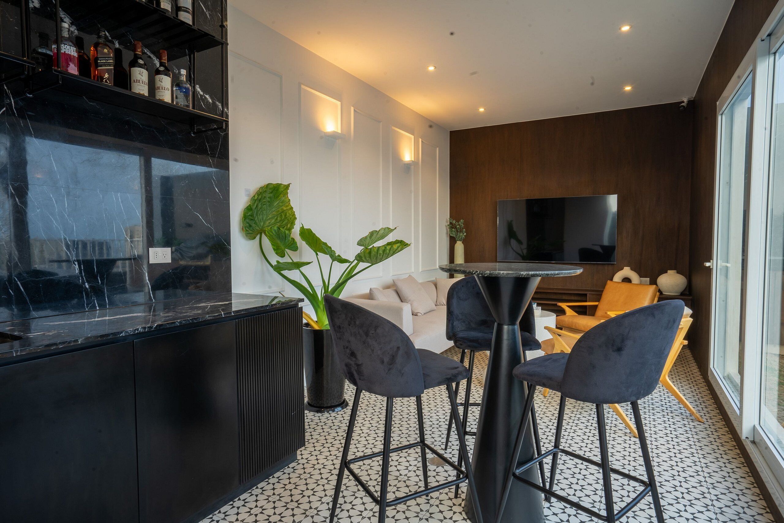 A modern kitchen with a marble countertop, a bar area with stools, a large potted plant, and a flat-screen TV in the background.