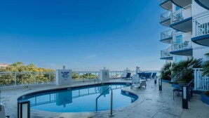 Una piscina en la azotea con vistas despejadas del horizonte urbano, rodeada de sillones de descanso y un edificio con balcones.