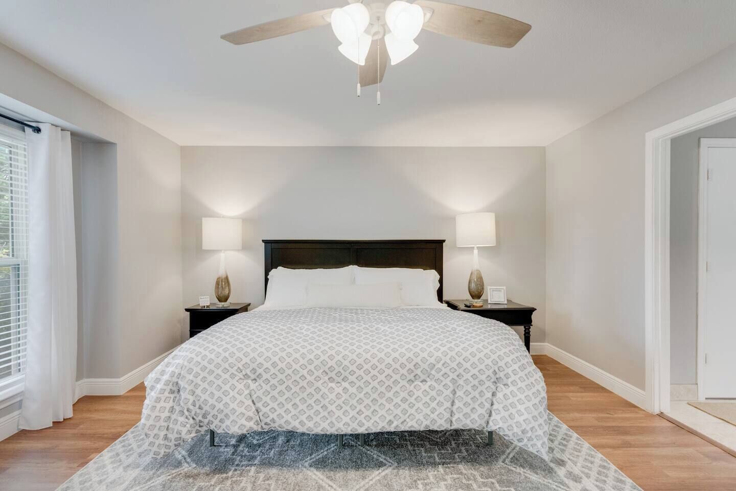 A bedroom with a bed, two bedside tables with lamps, a ceiling fan, and a window with curtains.