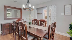 A dining room with a wooden table, six chairs, a chandelier, a mirror, and a potted plant.