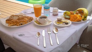 Aamiaistarjoilu uima-altaan äärellä: tarjolla on croissantteja, appelsiinimehua, kahvia ja hedelmiä.