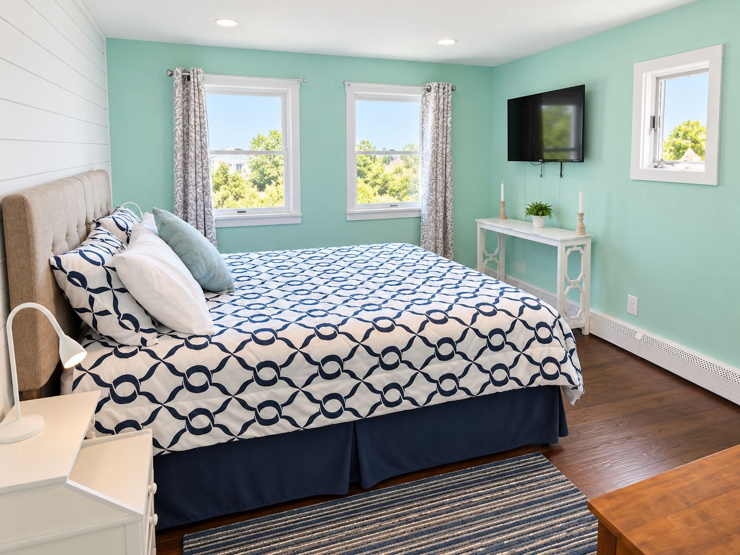 A bedroom with a bed, a bedside table, a TV, and two windows.