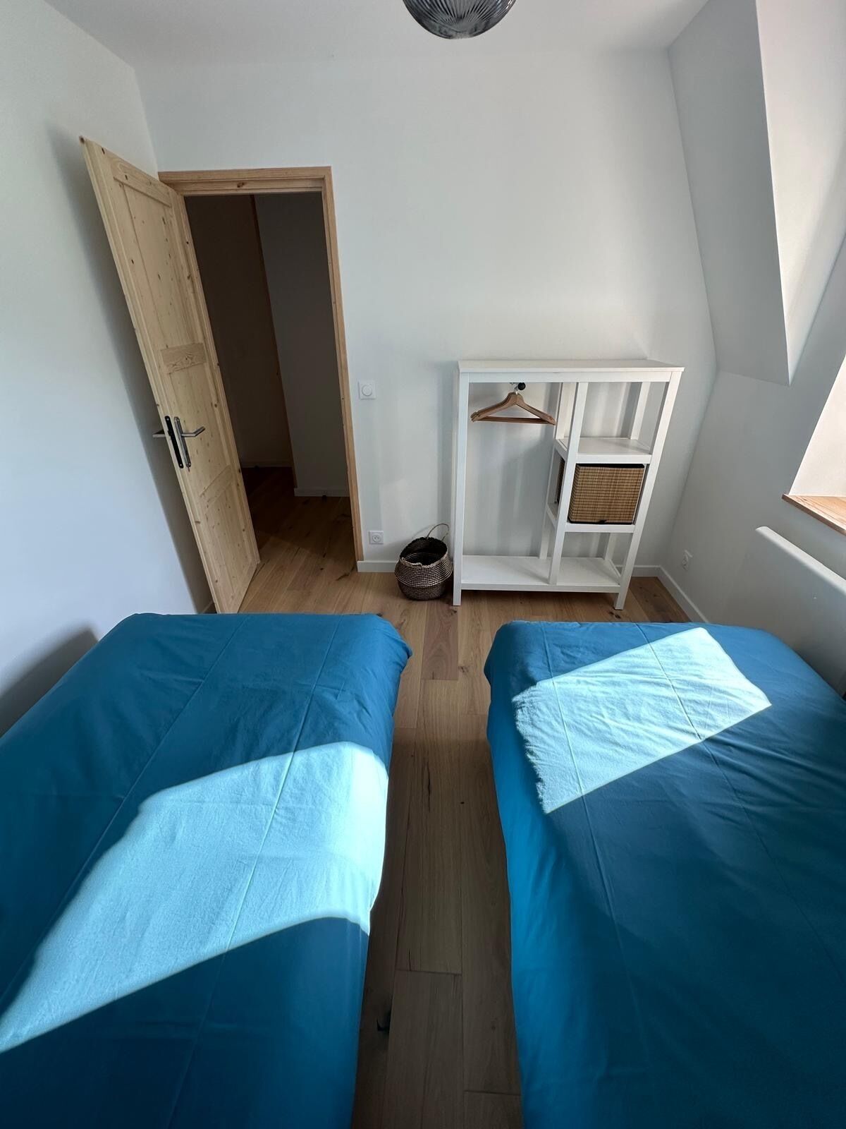 A room with two beds, a wardrobe, a hanging shelf, and a door leading to another room.