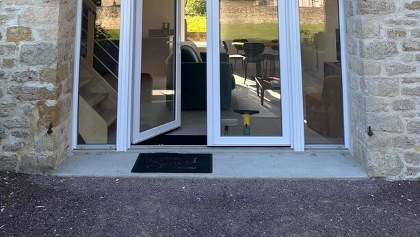 A stone building with a large arched entrance and a glass door. - Gîte 6 Personnes Chaleureux et au Calme à 15 Minutes des Plages du Débarquement (Picauville)