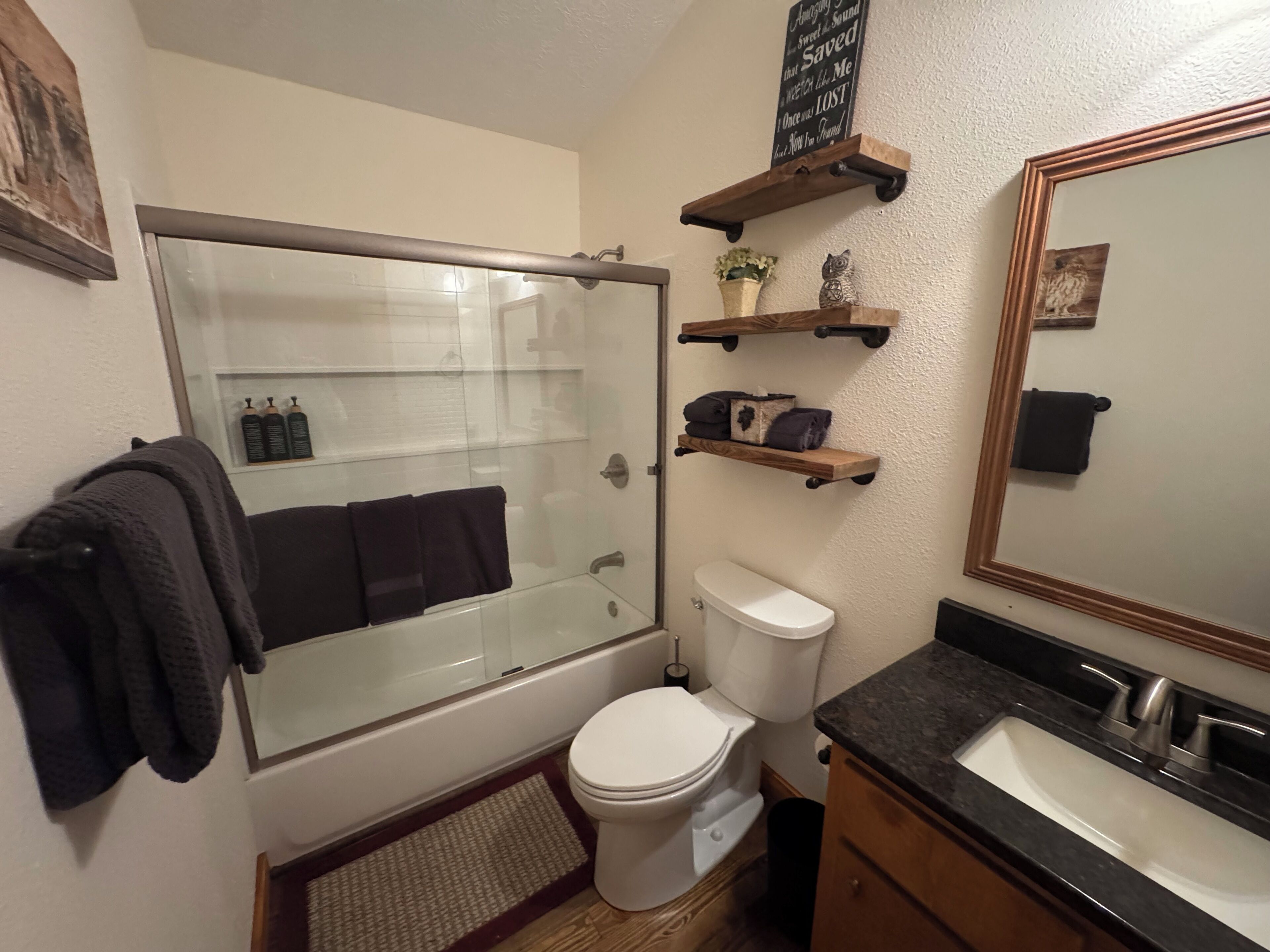 A bathroom with a glass-enclosed shower, a toilet, a sink, and wooden shelves above the toilet.