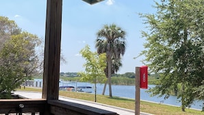 Una terraza de madera con baranda metálica, un espacio cubierto con techo verde, y vista a un lago rodeado de árboles y un buzón rojo.