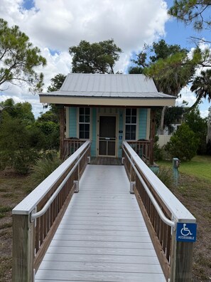 A building with a wooden deck leading to a door, surrounded by greenery. - Jiggs Landing, Cabin 2 - Bird Land Bunk House (Manatee County)