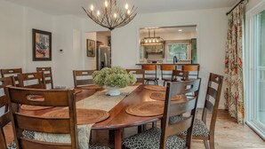 A dining area with a wooden table, chairs, a chandelier, and a kitchen in the background.