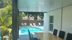 An outdoor patio with a pool, a covered dining area with a table and chairs, and a view of a backyard with a wooden fence and trees. - Pool entire house 3bed 2bath and sofa bed  (North Miami Beach)