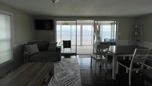 Ein Wohnzimmer mit einer Couch, einem Couchtisch und einem Fernseher. Es gibt einen Essbereich mit Tisch und Stühlen. Der Raum verfügt über eine große Schiebeglasdoor, die auf einen Balkon mit Blick aufs Meer führt.