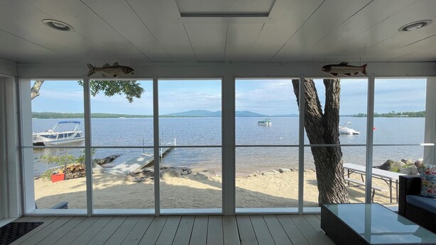 Ein Zimmer mit großen Fenstern, die auf einen See, eine Anlegestelle und einen Sandstrand blicken.