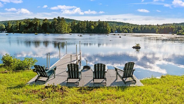 A dock with four Adirondack chairs facing a calm lake, surrounded by lush greenery and a forested hill in the background. - Lakefront Ski Log Cabin on 2 acres (Washington)