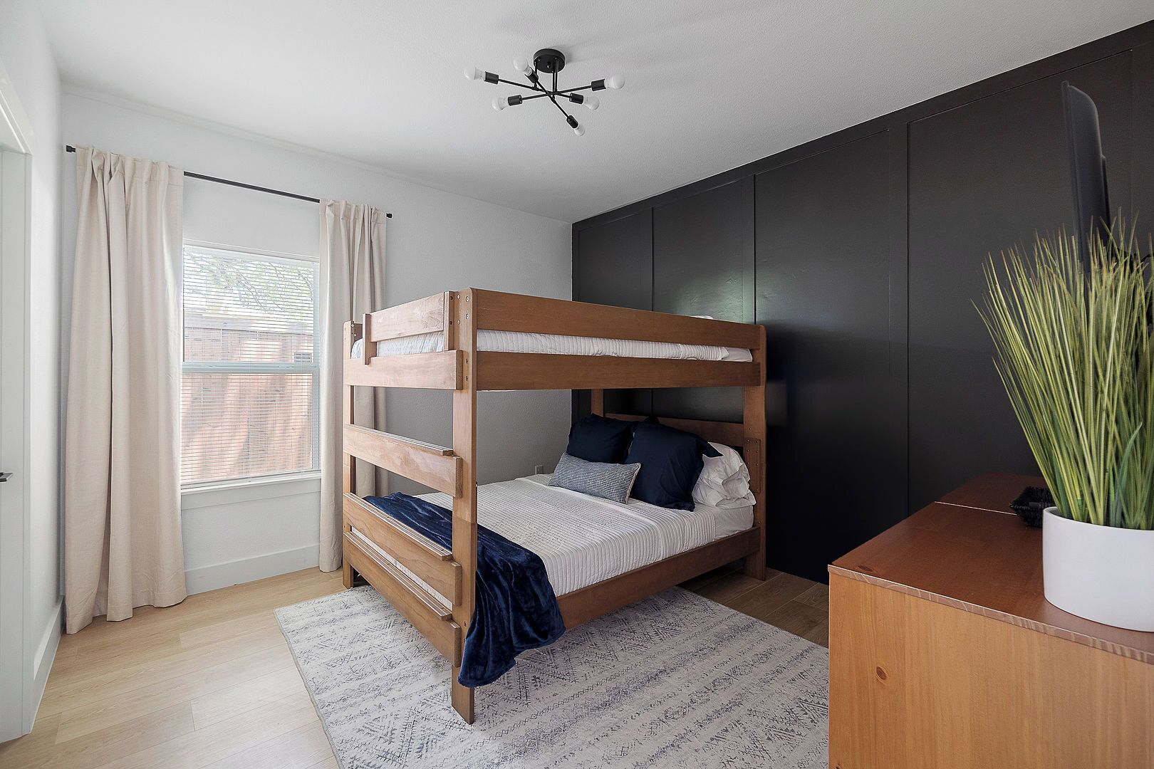 Una cama litera en una habitación con pared oscura, una ventana con cortinas y una lámpara en el techo.