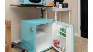 A compact kitchen setup with a blue microwave on a shelf, a small refrigerator with a freezer, and a wall clock. - Welcome to Casa Bugambilia  (Weslaco)