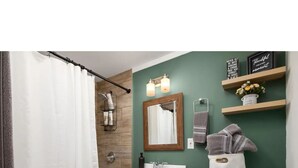 A bathroom with a shower curtain, a bathtub, a white sink with a mirror above it, and a toilet. There are shelves with decorative items and a towel rack. - Welcome to Casa Bugambilia  (Weslaco)