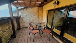 Een overdekt terras buiten, met een stenen muur, houten vloer en rieten stoelen rond een tafeltje.