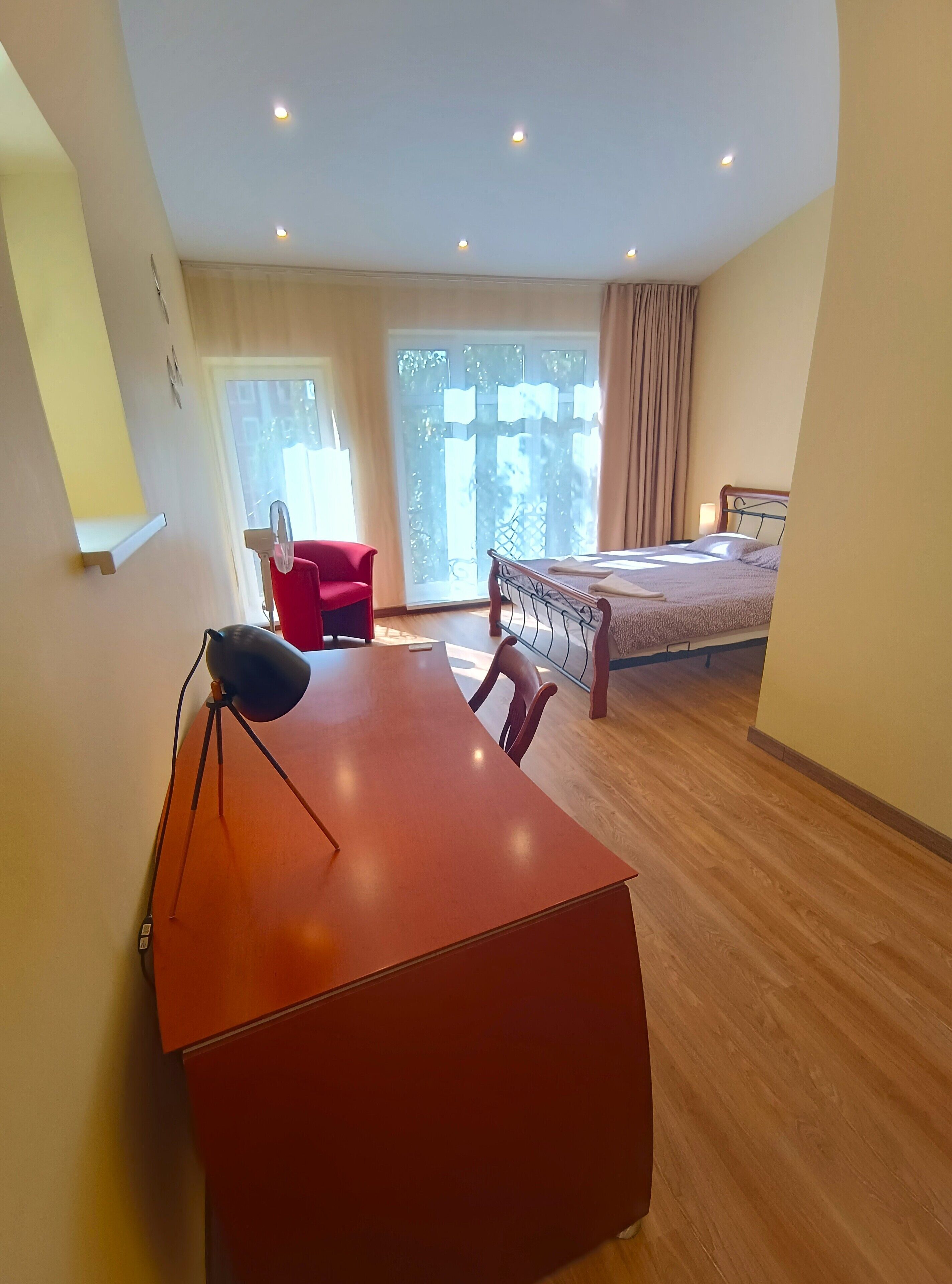 Une chambre d’hôtel moderne avec un plancher en bois, un lit avec un cadre en métal, une chaise rouge, un bureau avec une lampe et une grande fenêtre avec des rideaux.