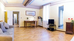 Een woonkamer met een houten visgraatvloer, een bank, een bureau met stoel, een televisie op een standaard en een radiator.