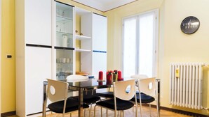 Een eethoek met een tafel en stoelen, een glazen kast met planken, en een klok aan de muur.